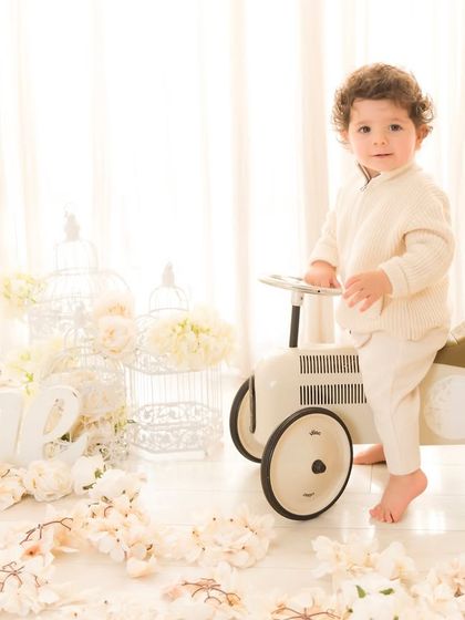 Ready for his first birthday photoshoot. This little boy looks dapper in his cream sweater, standing by a vintage-style toy car in a dreamy, all-white floral setup.