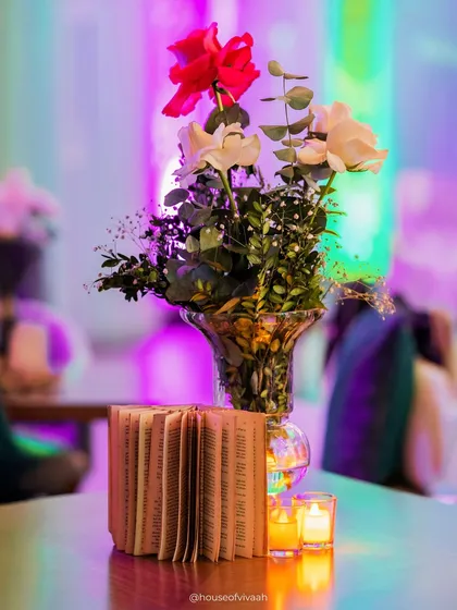 A simple yet evocative centerpiece featuring a book and a vase of roses, illuminated by candles against a backdrop of colorful party lights.