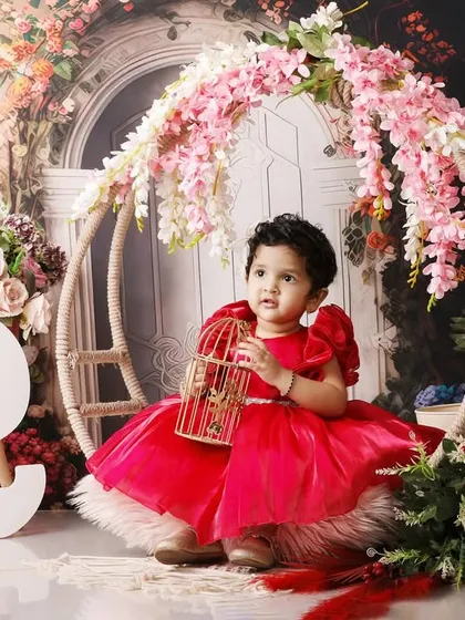 A little girl in a stunning red gown holding a birdcage, celebrating her second birthday in a floral-themed studio.