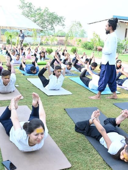 An outdoor class practicing Dhanurasana (Bow Pose) together on the grass. The fresh air and natural surroundings enhance the benefits of the practice.