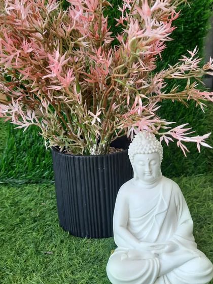 A simple yet powerful decor combination: a white meditating Buddha statue paired with a black planter holding pink artificial leaves. The contrast between the black, white, and pink is very striking.