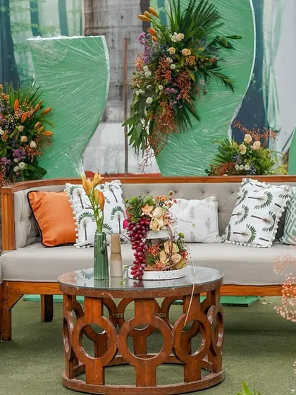 A comfortable lounge area with wooden furniture, set against a backdrop of green drapes and a forest print, with pops of orange and green in the cushions and florals.