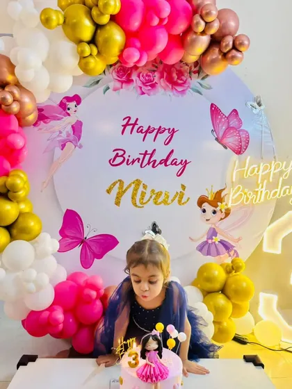 A candid moment from a fairy-themed 3rd birthday party. The birthday girl blows out her candles in front of a beautiful backdrop decorated with pink and gold balloons and fairy graphics.