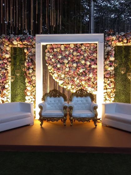 A creative and modern photo backdrop. A large white frame highlights a crescent of pink and white flowers against a green hedge, with elegant throne-like chairs for the couple.