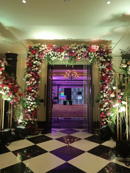 A grand floral entrance leading into a banquet hall with a striking black and white checkered floor. The abundance of pink, red, and white flowers creates a stunning contrast and a luxurious welcome.