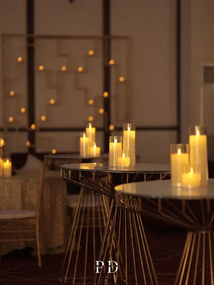 Elegant cocktail tables with clusters of candles, set against a softly lit backdrop. This creates intimate, warm spaces for guests to mingle during a Sangeet or reception.