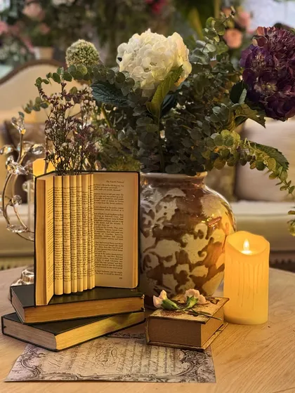 A table centerpiece that is a story in itself. A stack of vintage books, one artfully folded, is paired with a single candle and a floral arrangement, creating a poetic and literary-inspired detail.