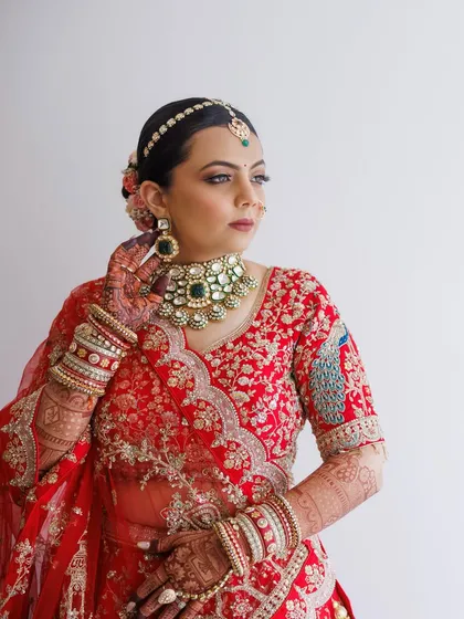 A confident pose showcasing the stunning emerald jewelry against the red lehenga. The makeup is perfectly balanced to complement the rich colors of her attire.