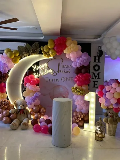 A chic and modern first birthday setup for baby Shrivi. The design features a glowing crescent moon prop, a 'Happy Birthday' neon sign, and a beautiful organic balloon arch in pastel and metallic tones, complete with a teddy bear.