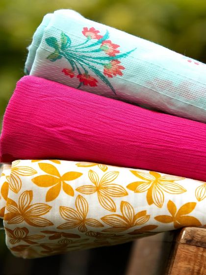 A creative shot of three rolled-up fabrics with different prints and textures, stacked on a wooden surface outdoors to show them in natural light.