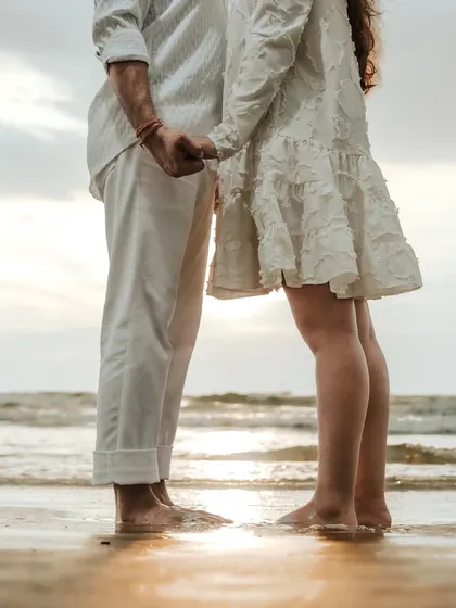 A close-up, artistic shot of a couple holding hands at the water's edge. The focus on their feet in the sand and the gentle waves creates a feeling of intimacy and connection.
