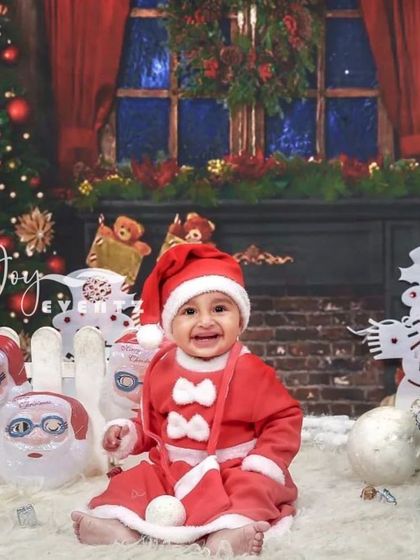 A festive Christmas-themed photoshoot to capture the holiday magic. This baby boy, dressed in a Santa suit, smiles happily in a cozy winter wonderland setup complete with a tree and snowman.