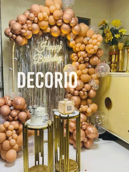 A glamorous birthday setup featuring a full arch of rose gold balloons against a silver fringe backdrop. The decor is completed with elegant gold pedestals for the cake and gifts.