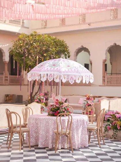 An elegant table setting at the Jaipur welcome lunch. A custom-printed umbrella provides shade over a table with a patterned cloth and a beautiful floral centerpiece, creating a chic and inviting dining spot.