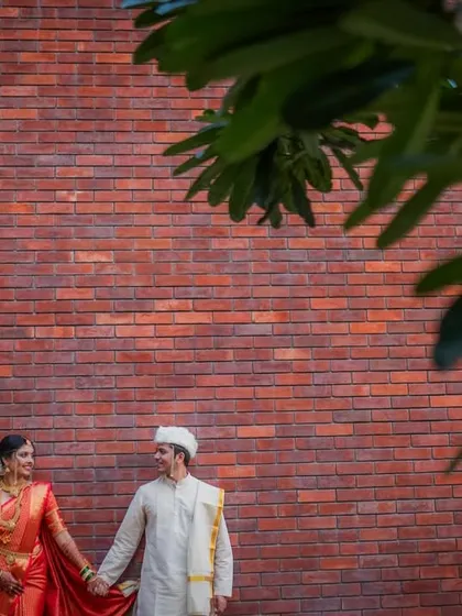 The couple holds hands, standing against a striking red brick wall. The artistic framing with a tree branch adds a touch of nature to this modern-meets-traditional portrait.