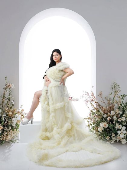 A full-length view of the stunning cream-colored ruffled gown. The mom-to-be is posed elegantly on a platform, showing off the dress's long, beautiful train.