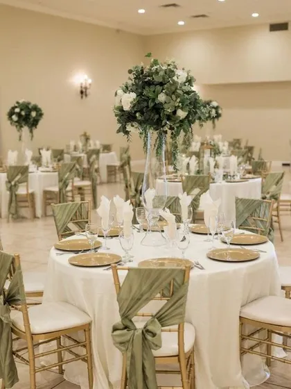 An indoor reception hall with round tables and Chiavari chairs decorated with sage green sashes. The tall floral centerpieces add height and drama to the room.