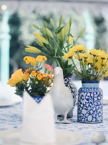 A fun 'Masakali' inspired tablescape for a Haldi lunch. We used blue pottery vases, bright yellow flowers, and a printed tablecloth to create a cheerful setting, with a little white pigeon prop adding a playful touch.