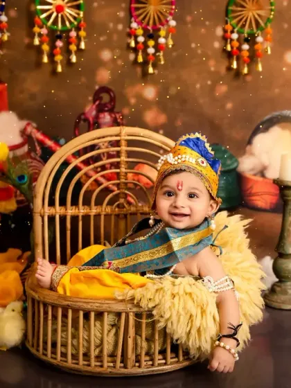 A wide shot of the elaborate Little Krishna setup, showing the baby smiling in his wicker chair surrounded by festive Indian decorations.