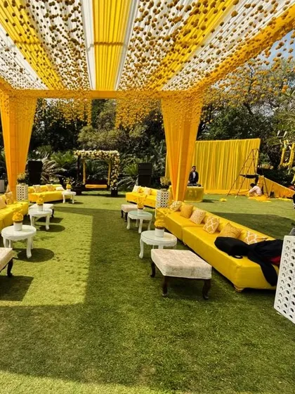 A wide view of a lawn decorated for a Haldi ceremony. The space is filled with yellow lounge sofas and a canopy of white and yellow drapes with hanging marigold balls.