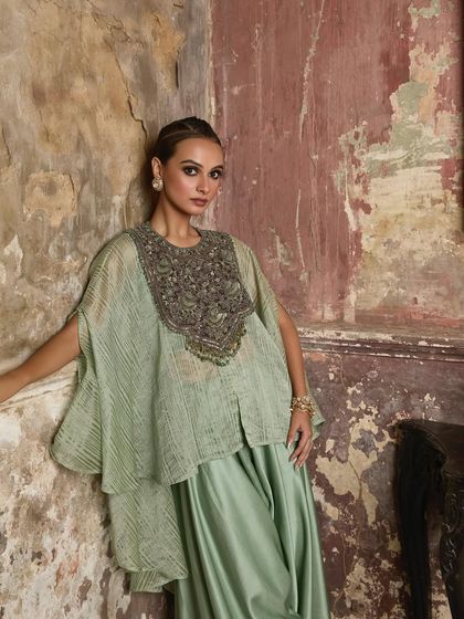 A model in a pastel green layered cape set, the textures of her outfit beautifully complementing the rustic backdrop.