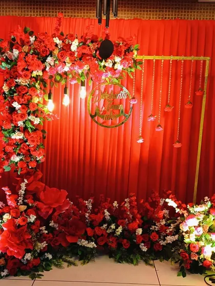 A full view of a stunning red floral backdrop, perfect for an anniversary or a romantic birthday celebration. The design includes a custom gold nameplate and hanging Edison bulbs.