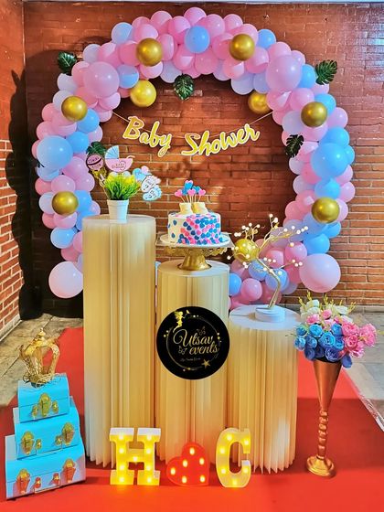 A baby shower decoration with a pink and blue balloon arch against a brick wall. The setup includes plinths for the cake and other decorative items.