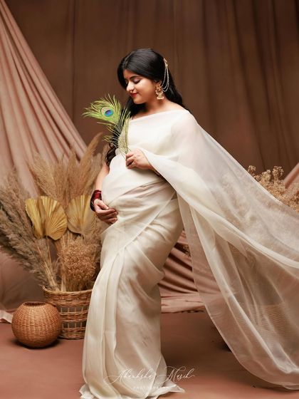 A beautiful side profile in a white saree, set against a rustic backdrop. The peacock feather adds a touch of color and traditional symbolism.