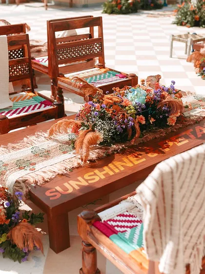 A rustic-chic seating arrangement with woven chairs and a wooden bench inscribed with "Sunshine & Petals". Personalization is key to making an event feel special.