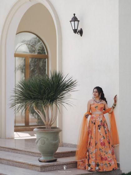 A full-length portrait of the bride in her orange floral lehenga, posing gracefully before her Haldi ceremony.