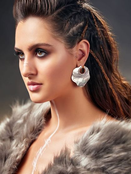 A close-up beauty shot from the Pure Facets campaign, highlighting the unique hammered-metal earring and fierce makeup.