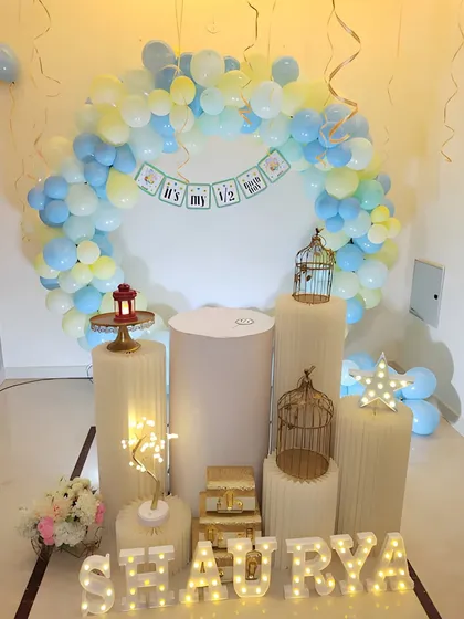 An elegant half-birthday celebration corner. This setup uses a circular balloon arch in pastel blue and yellow, illuminated marquee letters for the baby's name, and decorative props like plinths and birdcages.