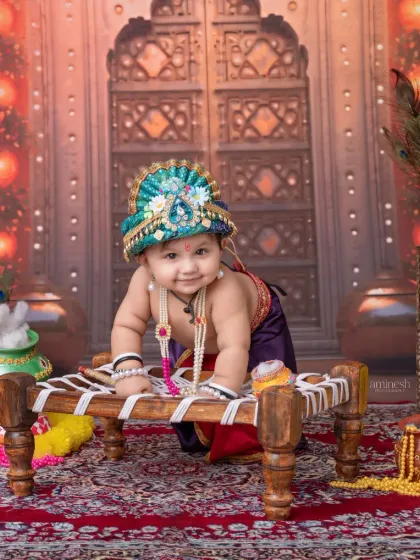 This little Krishna is ready to play, captured in a dynamic pose on a small wooden cot. The composition balances the child's energy with the ornate, traditional backdrop.