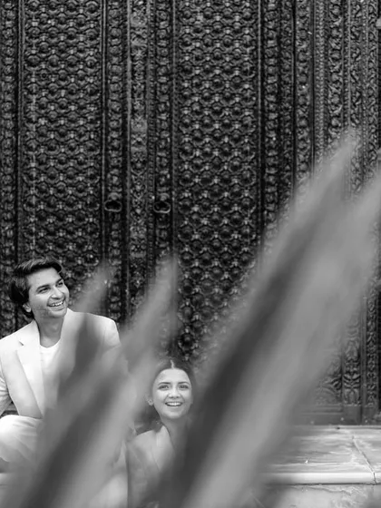 A creative composition, shooting through foreground foliage to capture the couple's happy expressions. This black and white shot adds depth and focuses on their joyful interaction in front of a traditional carved door.
