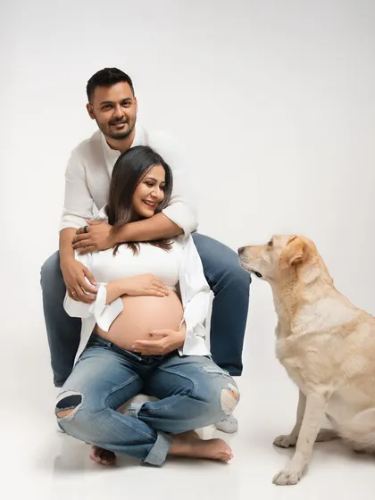 A classic maternity pose with the couple and their dog looking on. The father-to-be has his arms wrapped around the mother, creating a feeling of love and protection.