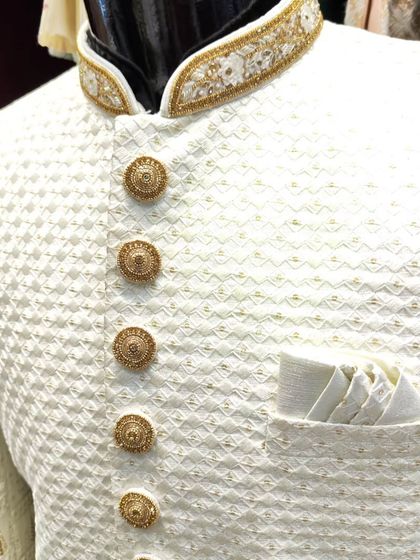 A close-up of a white textured sherwani, showing the intricate weave of the fabric, the embroidered collar, and the elegant gold buttons.