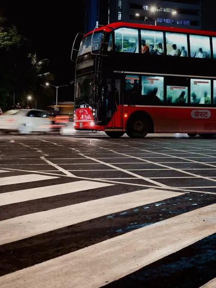 A classic red double-decker BEST bus crosses a zebra crossing in Mumbai at night. The long exposure shot captures the motion of a passing car as a white streak, contrasting with the stationary bus.