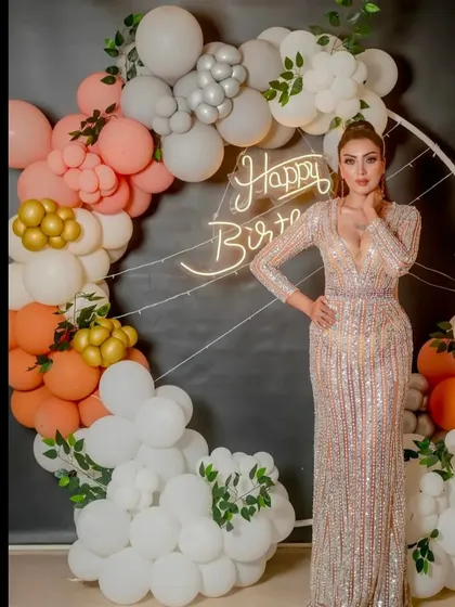 A glamorous birthday girl posing in front of a custom balloon installation. The organic balloon arrangement in peach, white, and gold with hints of greenery perfectly complements her sparkling dress.