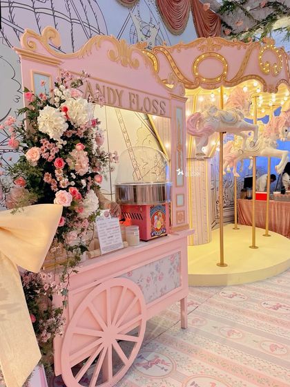 A vintage-style candy floss cart, adorned with flowers, stands next to the magical carousel.