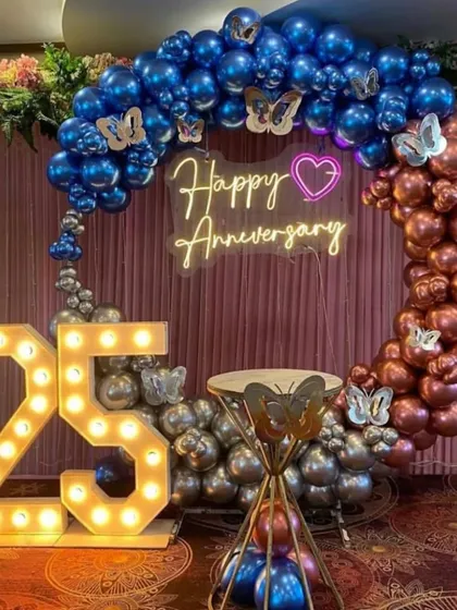 A glamorous 25th-anniversary party with a two-toned balloon ring in chrome blue and rose gold. The setup includes large marquee numbers and a "Happy Anniversary" neon sign.