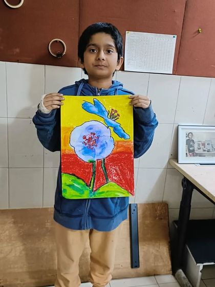The finished masterpiece! A student proudly holds up his completed painting of a blue flower with a dragonfly, a testament to his hard work.