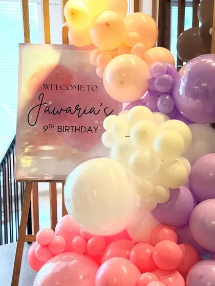 A personalized welcome sign for "Jawaria's 9th Birthday" on an easel, adorned with a beautiful arrangement of pink, purple, and white balloons. The watercolor-style sign adds a touch of artistic flair to the party entrance.
