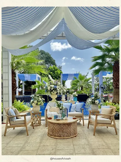 A poolside lounge setup for a daytime brunch. The airy blue and white striped drapes, natural cane furniture, and fresh floral arrangements create a relaxed, sophisticated atmosphere that is perfect for a sunlit celebration.