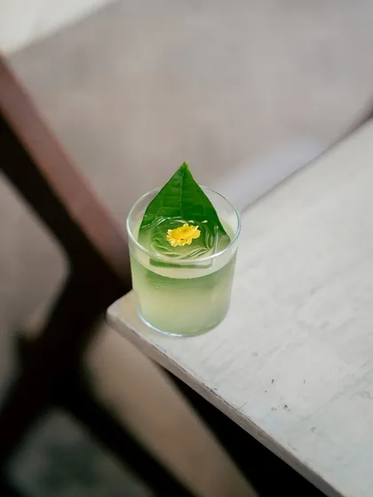 A close-up of the Queen's Kiss, where a vibrant yellow flower rests on a kaffir lime leaf. This cocktail blends rum and exotic spices, creating a drink that is as fragrant and enchanting as its namesake, perfect for a royal moment.