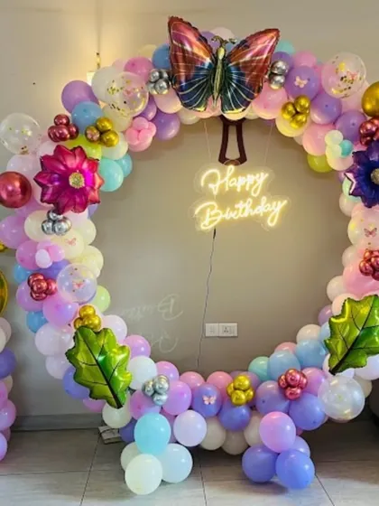 This colorful butterfly-themed decoration features a full ring of pastel balloons. The design is accented with butterfly and leaf foil balloons, a large number '5' foil balloon, and a central 'Happy Birthday' neon sign.