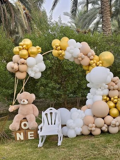 A beautiful outdoor first birthday party with a teddy bear theme. The decor features a large organic balloon arch in neutral tones, a light-up number '1', and a cute teddy bear holding balloons.