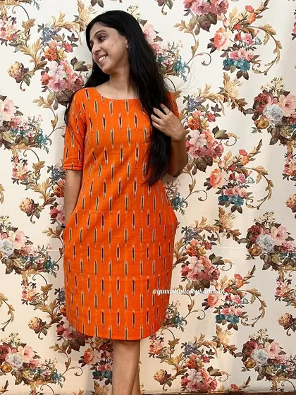 A full view of the orange straight-fit Ikat dress against a floral background. It’s a simple, elegant, and affordable piece from my handloom collection.