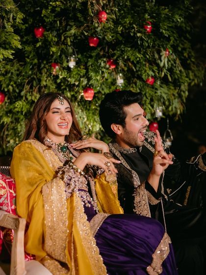 A candid moment of laughter and joy during the Mehfil-e-Mehendi. The couple is seated in front of a custom-designed apple tree, a key element of "The Perfumed Orchard" theme.