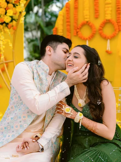 A tender moment from a Haldi ceremony, where the groom gently kisses his bride's cheek. A sweet and candid expression of love.