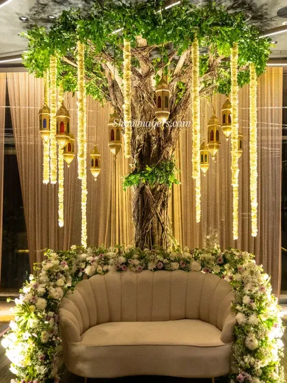We create serene and beautiful spaces for traditional ceremonies. This seating area, designed for a Mehendi, features an enchanting 'tree of life' centerpiece with hanging floral strings and lanterns, set against a backdrop of elegant curtains, providing a perfect photo opportunity.
